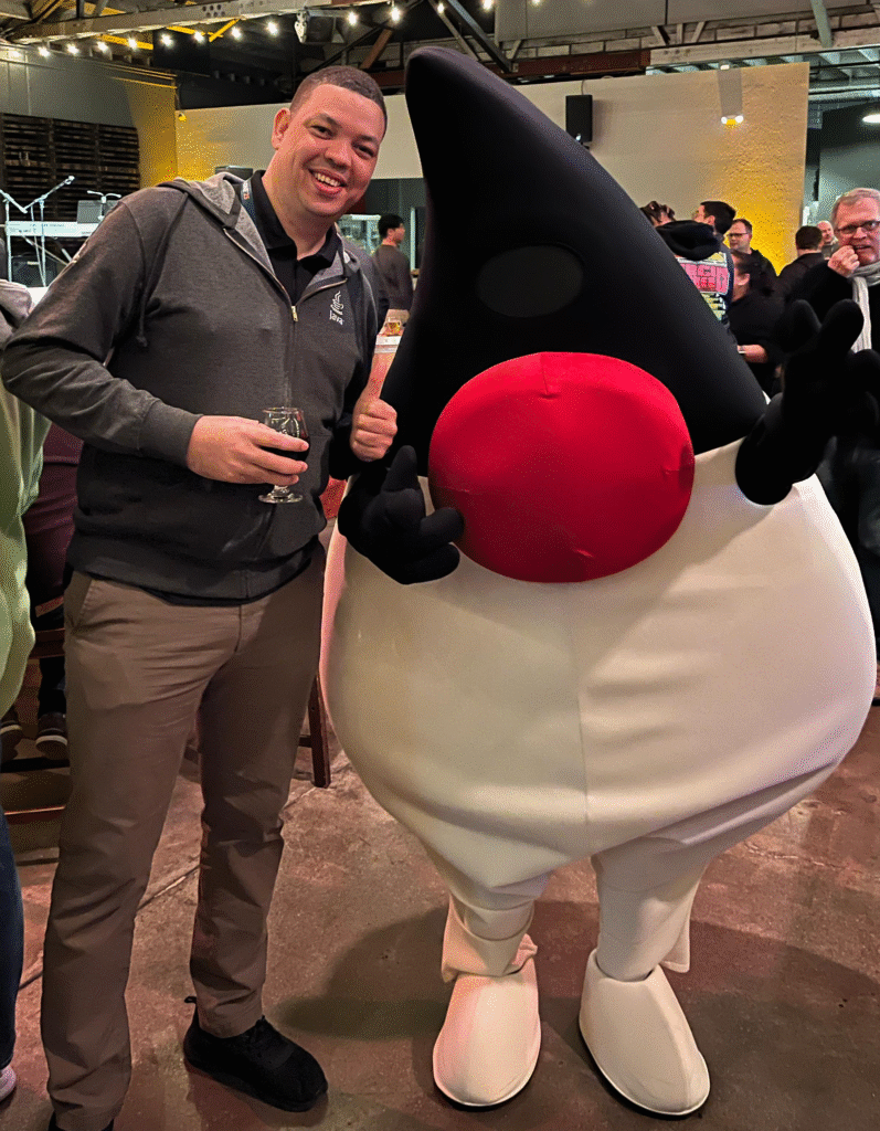 A smiling man stands indoors next to a person dressed in a large Duke the Java mascot costume. The man is holding a drink and posing with one arm around the mascot. The background shows people socialising in a warmly lit event space.