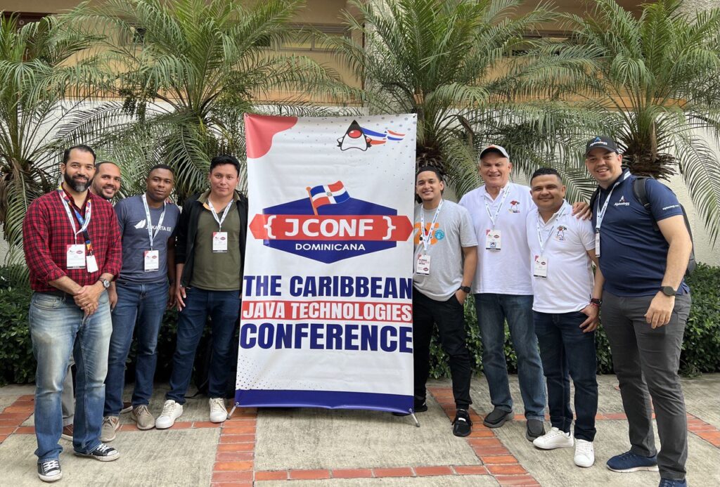 A group of nine open source enthusiasts standing outdoors in front of palm trees, smiling at the camera. They are gathered around a large banner that reads “JCONF Dominicana – The Caribbean Java Technologies Conference.” The group appears to be conference attendees or organisers, wearing name badges and casual clothing.