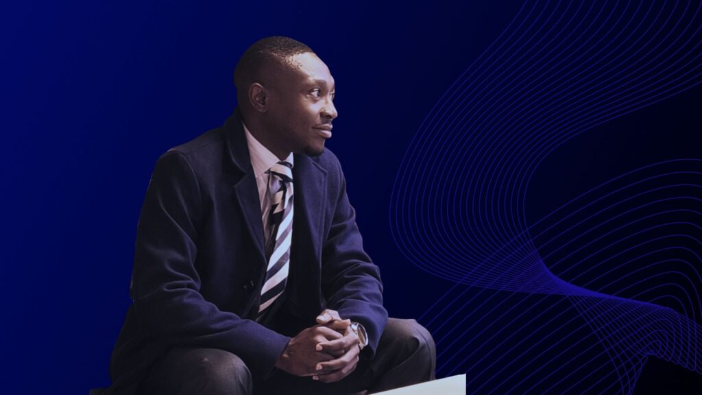 A man in a dark coat, dress shirt, and striped tie sits on a brown tufted leather sofa. He is turned slightly to the side with his hands clasped, looking thoughtfully into the distance. Behind him is a large mural depicting historical figures in a village scene.