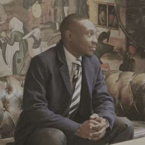 A well-dressed man sits on a tufted leather sofa, wearing a dark coat, dress shirt, and striped tie. He is turned slightly to the right with his hands clasped, appearing thoughtful. Behind him is a detailed, mural-style painting depicting historical figures in muted tones.