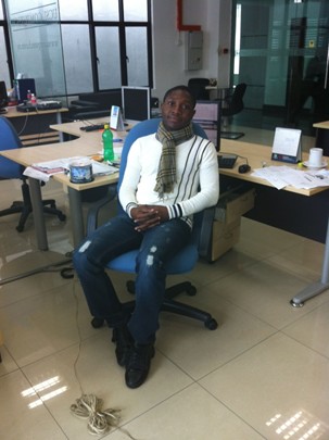 A man sits in a blue office chair in a modern office space. He is wearing a white sweater, a striped scarf, and ripped jeans. His hands are folded in his lap, and he faces the camera. Around him are desks with computers, papers, and office supplies. A coiled cord lies on the floor near his feet.