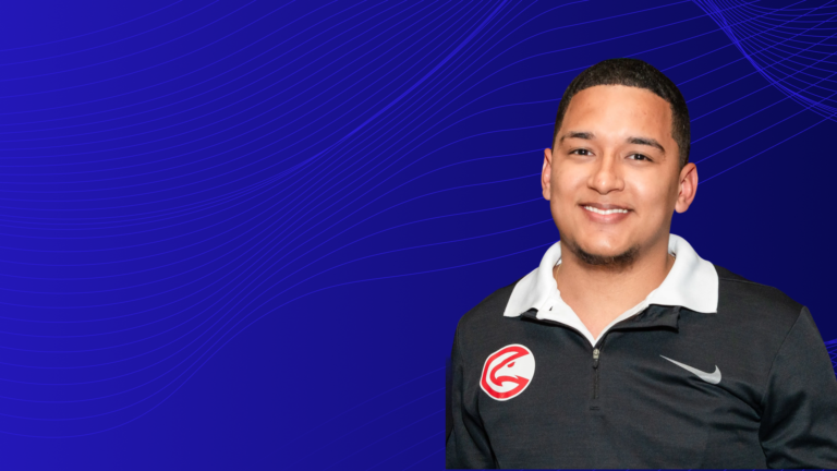 Smiling man wearing a black quarter-zip athletic top with a white collar and a red bird logo on the chest, standing against a blue abstract background with flowing wave lines.