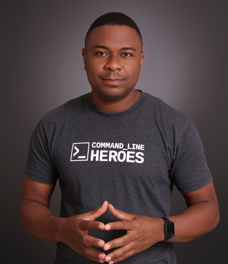 A man standing against a plain dark background, wearing a gray “Command Line Heroes” T-shirt. He has short hair, a neutral expression, and is holding his hands together in front of him. He is also wearing a smartwatch on his left wrist.