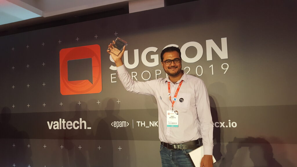 Man wearing a white shirt, glasses, and conference lanyard standing in front of a logo, holding his right hand up