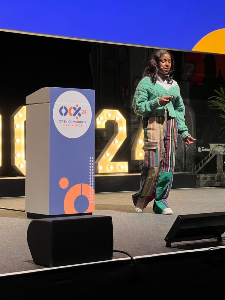 A woman stands on a conference stage giving a presentation, holding a clicker and speaking beside a podium labeled “OCX24 Open Community Experience.” She wears a green sweater and colorful patchwork pants. Large illuminated numbers “24” and stage lighting form the backdrop.