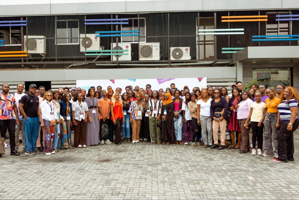 A large group of people stand together outdoors in front of a building, posing for a group photo. Many wear name badges and casual or business-casual clothing, suggesting a conference or community event.