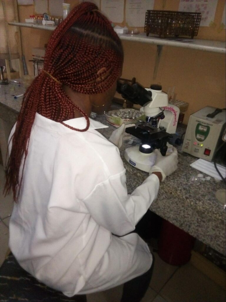A person wearing a white lab coat and gloves sits at a laboratory bench, looking through a microscope. They have long, red braided hair tied back. The workspace includes test tubes, sample containers, and lab equipment arranged on shelves and the countertop.