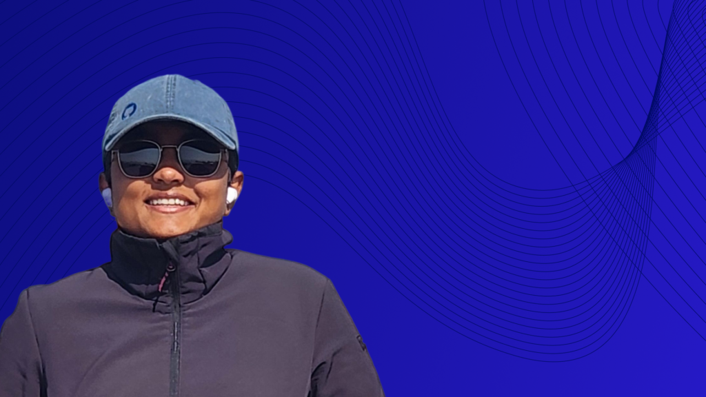 Portrait of a smiling person wearing a dark jacket, sunglasses, wireless earbuds, and a gray baseball cap against a blue abstract background with flowing line patterns.