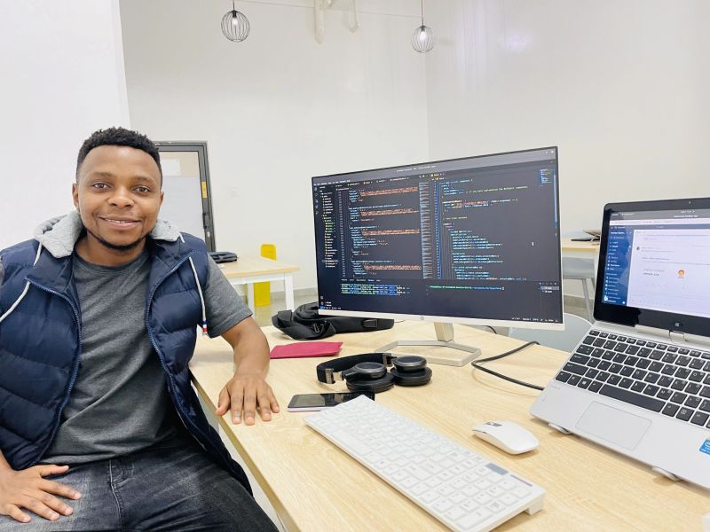Young man sitting at a desk with a laptop, a keyboard, and a monitor, looking at the camera