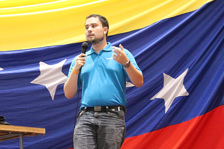 A man with microphone and blue t-shirt standing in front of a Venezuelan flag